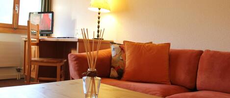 Una sala de estar acogedora con una mesa de madera, un sofá con cojines naranja, una lámpara y un televisor sobre un mueble de madera.
