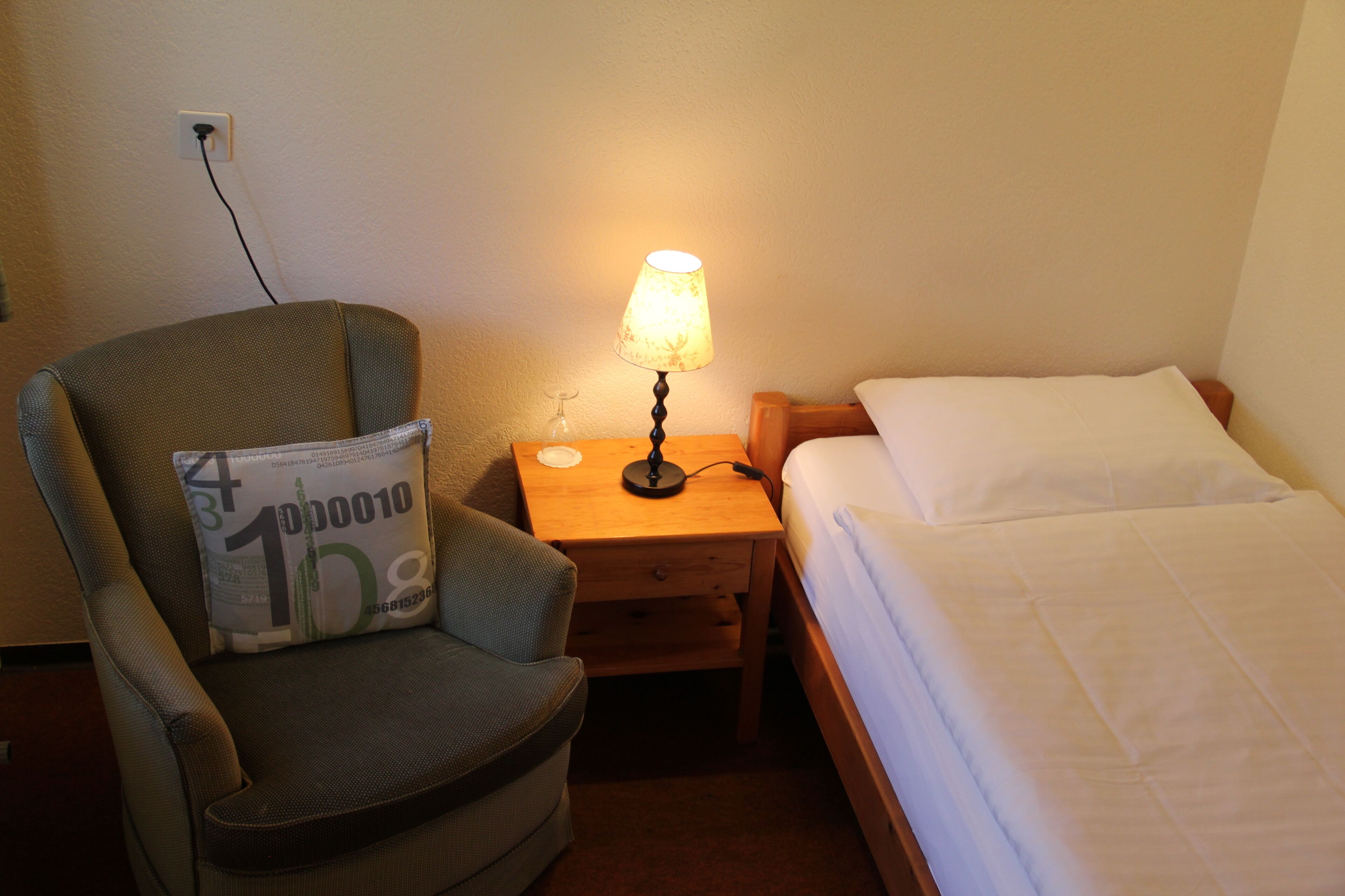 A hotel room with a single bed, a chair with a pillow, a nightstand with a lamp, and a power outlet on the wall.