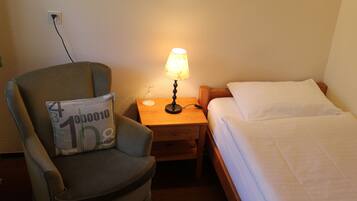 A hotel room with a single bed, a chair with a pillow, a nightstand with a lamp, and a power outlet on the wall.