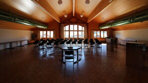 Un salón amplio con techo y paredes de madera, varias filas de sillas y ventanas grandes.