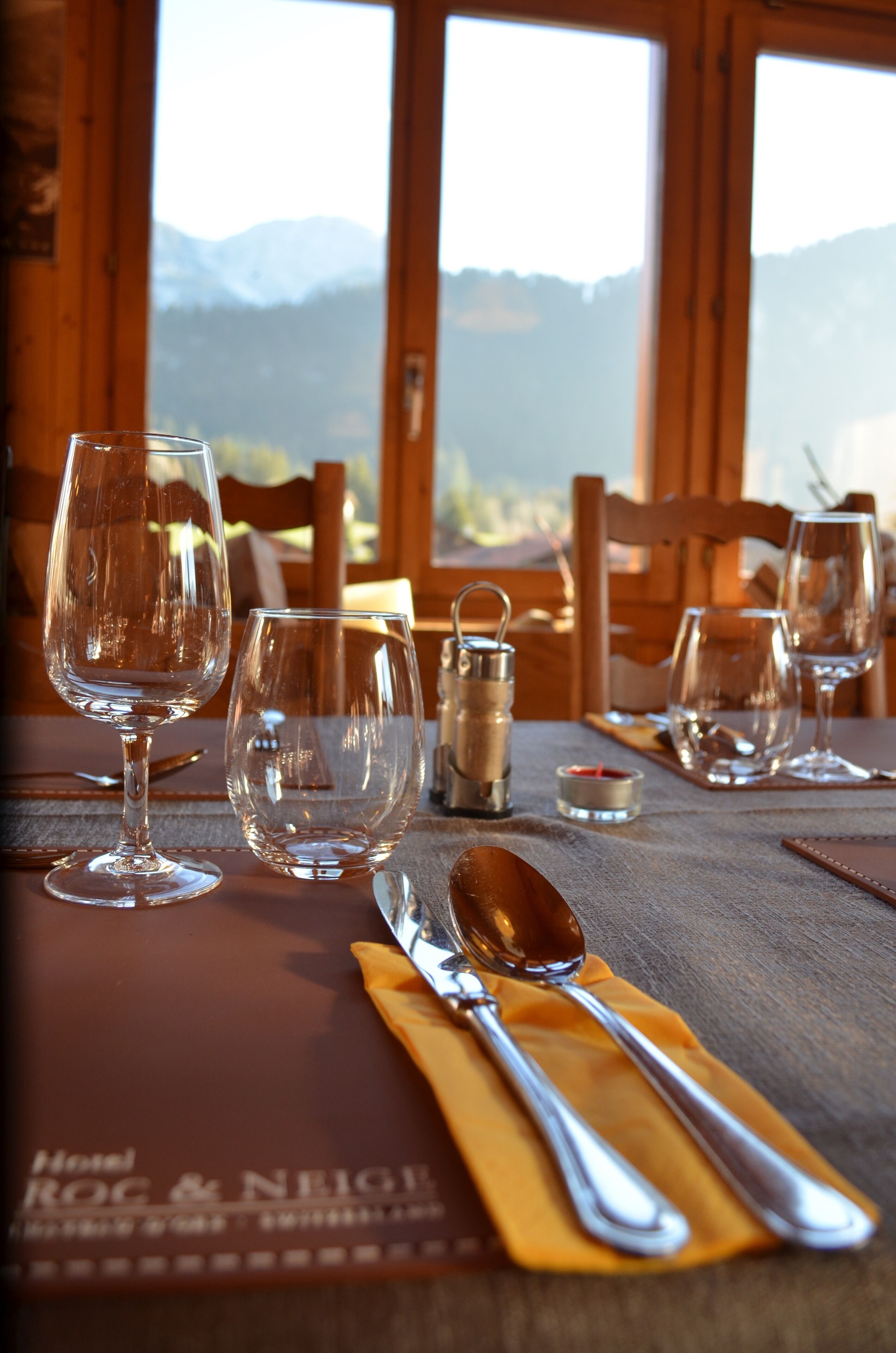 Une table à manger dressée avec des verres à vin, une bougie et des couverts, offrant une vue sur les montagnes à travers de grandes fenêtres.