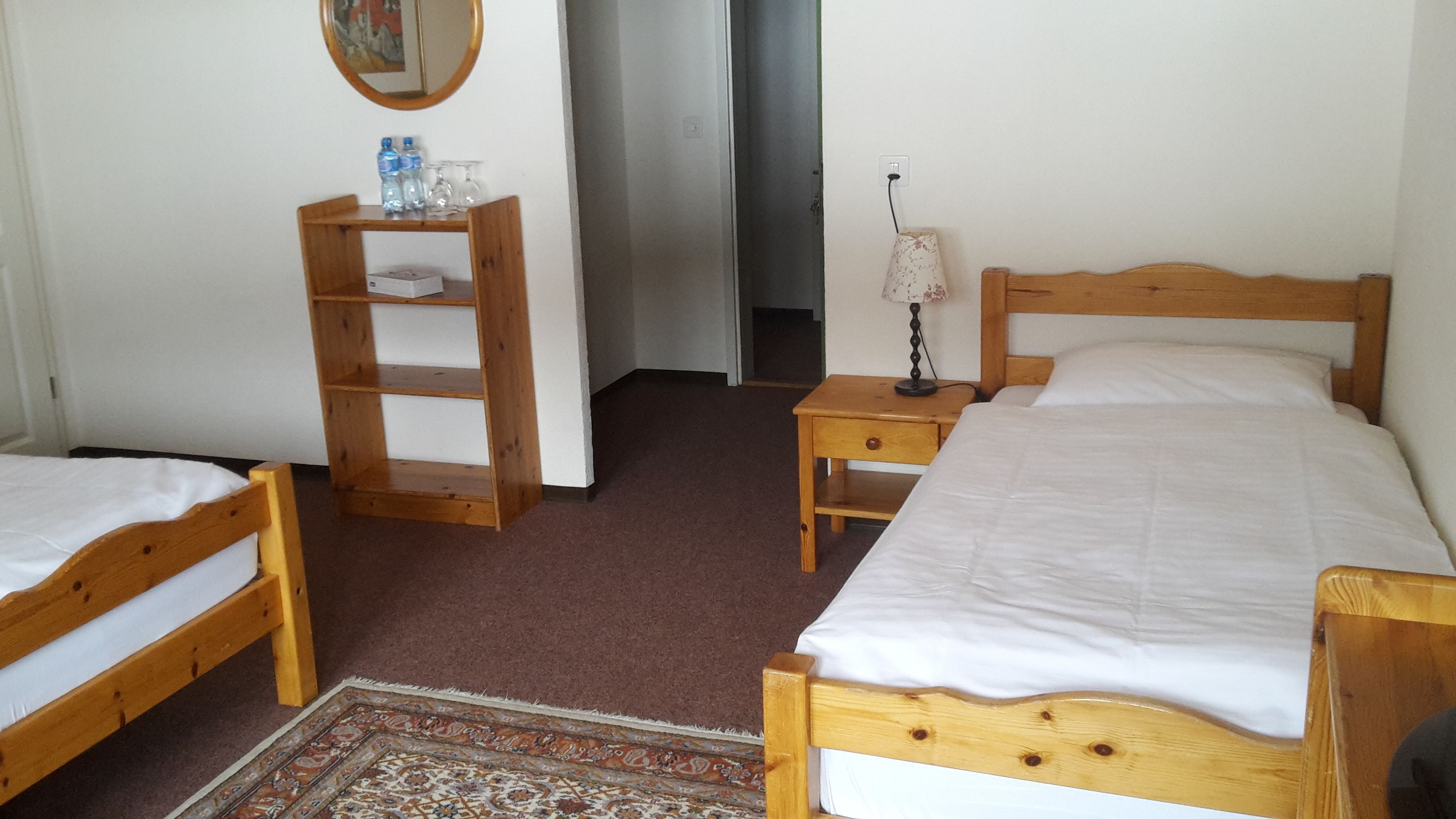 A bedroom with two wooden beds, a small wooden shelf with water bottles and glasses, a nightstand with a lamp, and a patterned rug on the floor.