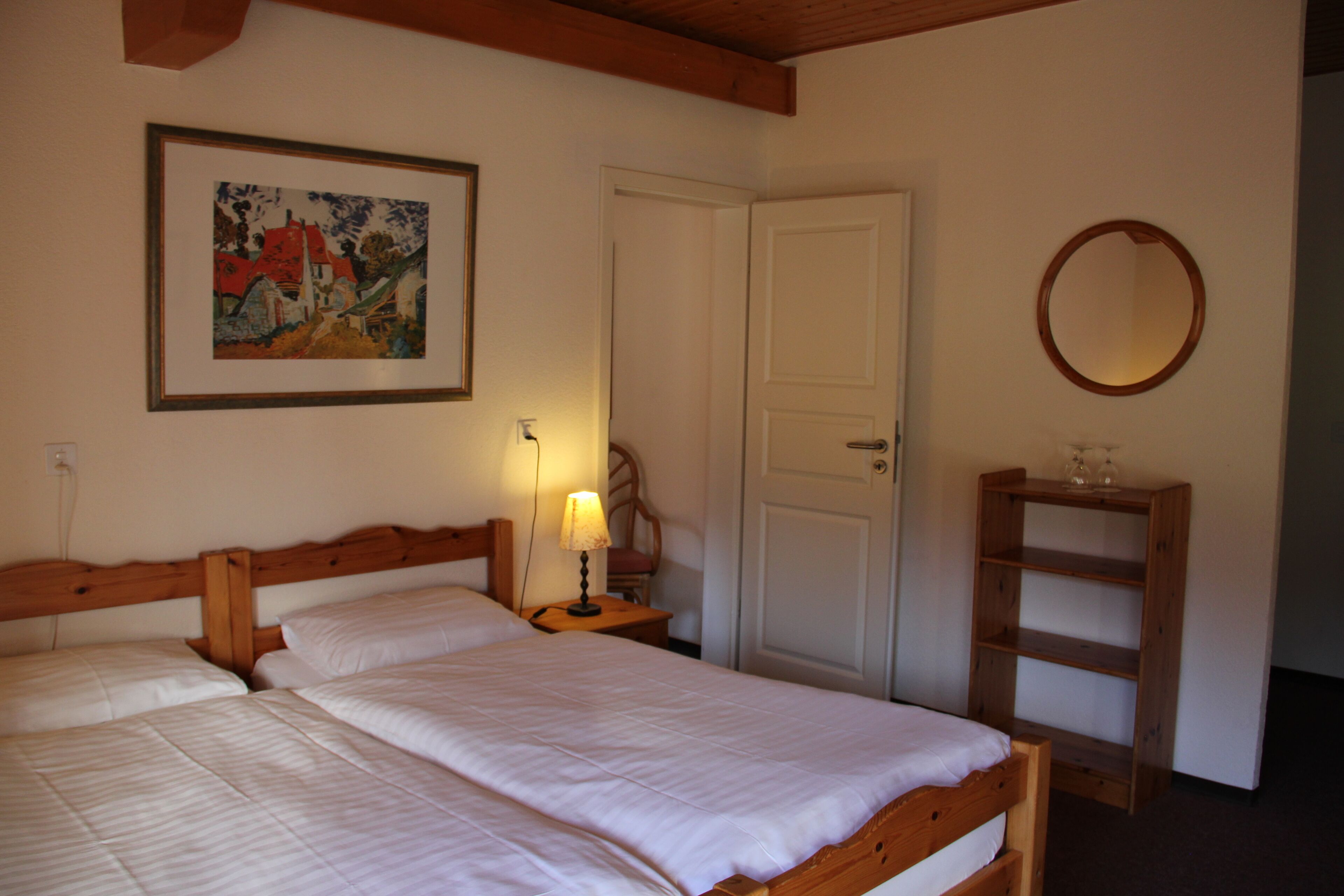 Un dormitorio con dos camas, cabeceros de madera, una mesita de noche con lámpara, una puerta, un espejo y una repisa con vasos.