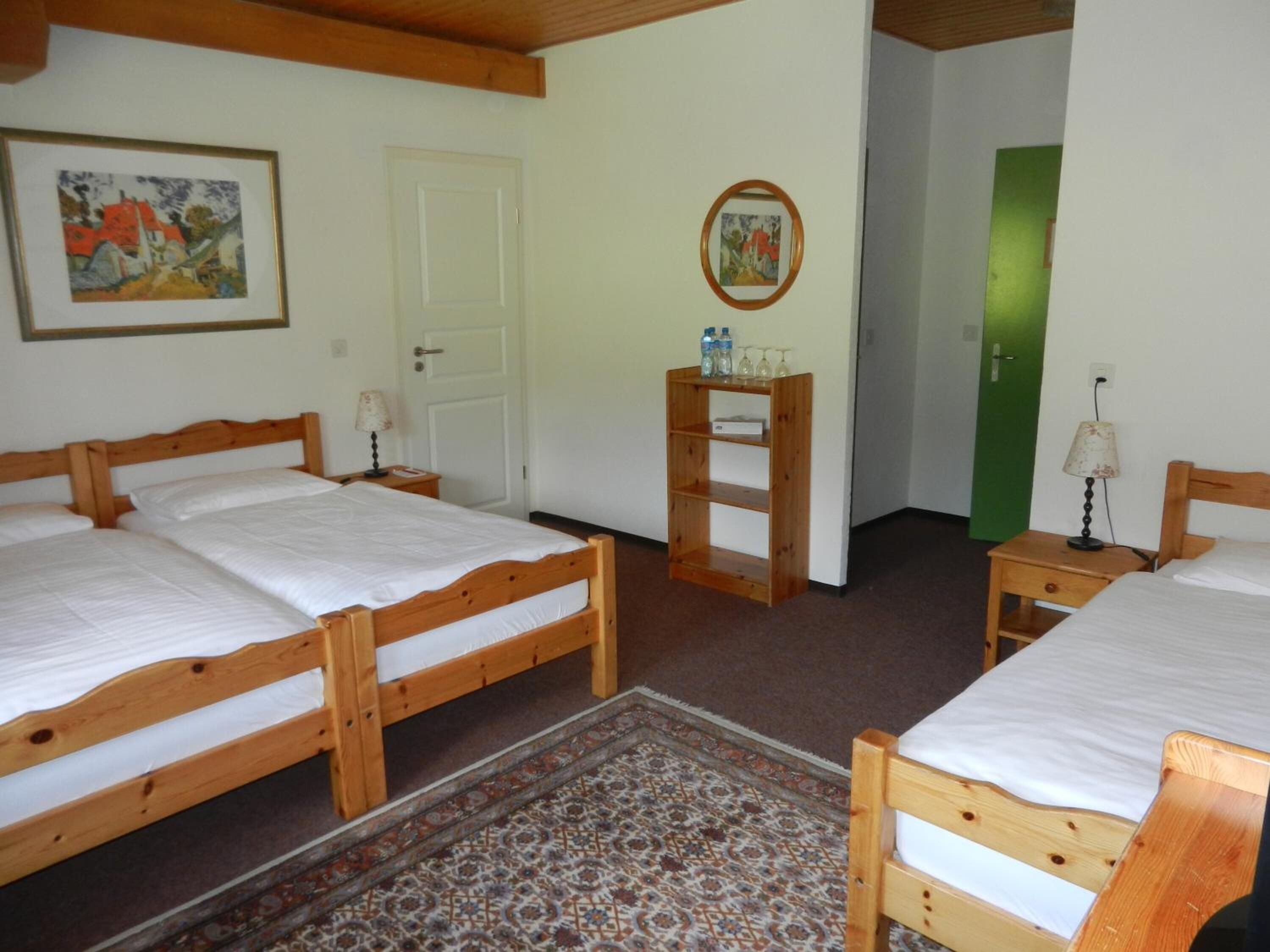 A room with two wooden beds, a small wooden nightstand with a lamp, a framed picture on the wall, and a door with a green frame.