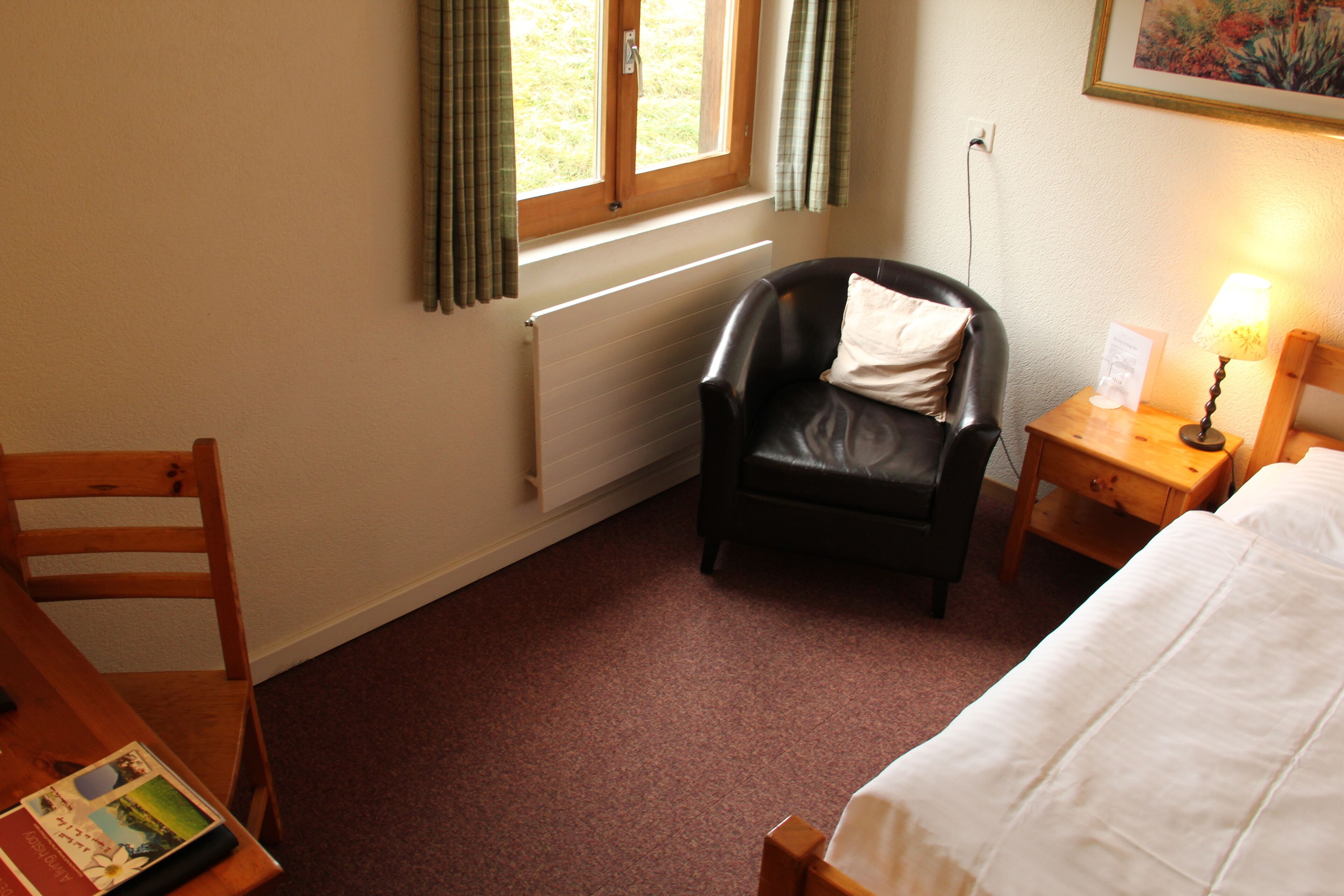 A hotel room with a bed, a chair, a table with a book, a lamp, a window with curtains, and a radiator.