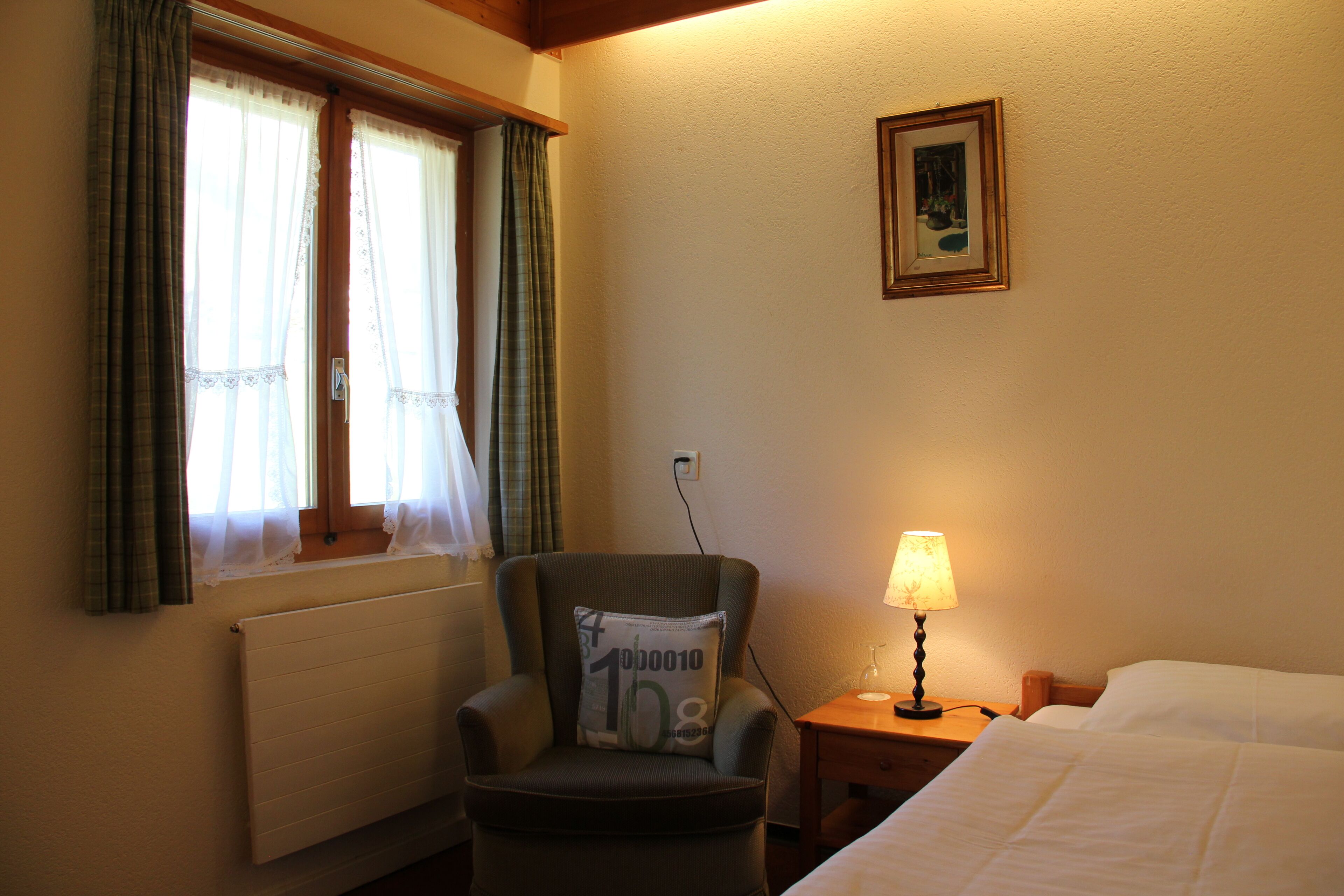 Habitación de hotel con cama, sillón con cojín, mesita de noche con lámpara, ventana con cortinas e imagen colgada en la pared.