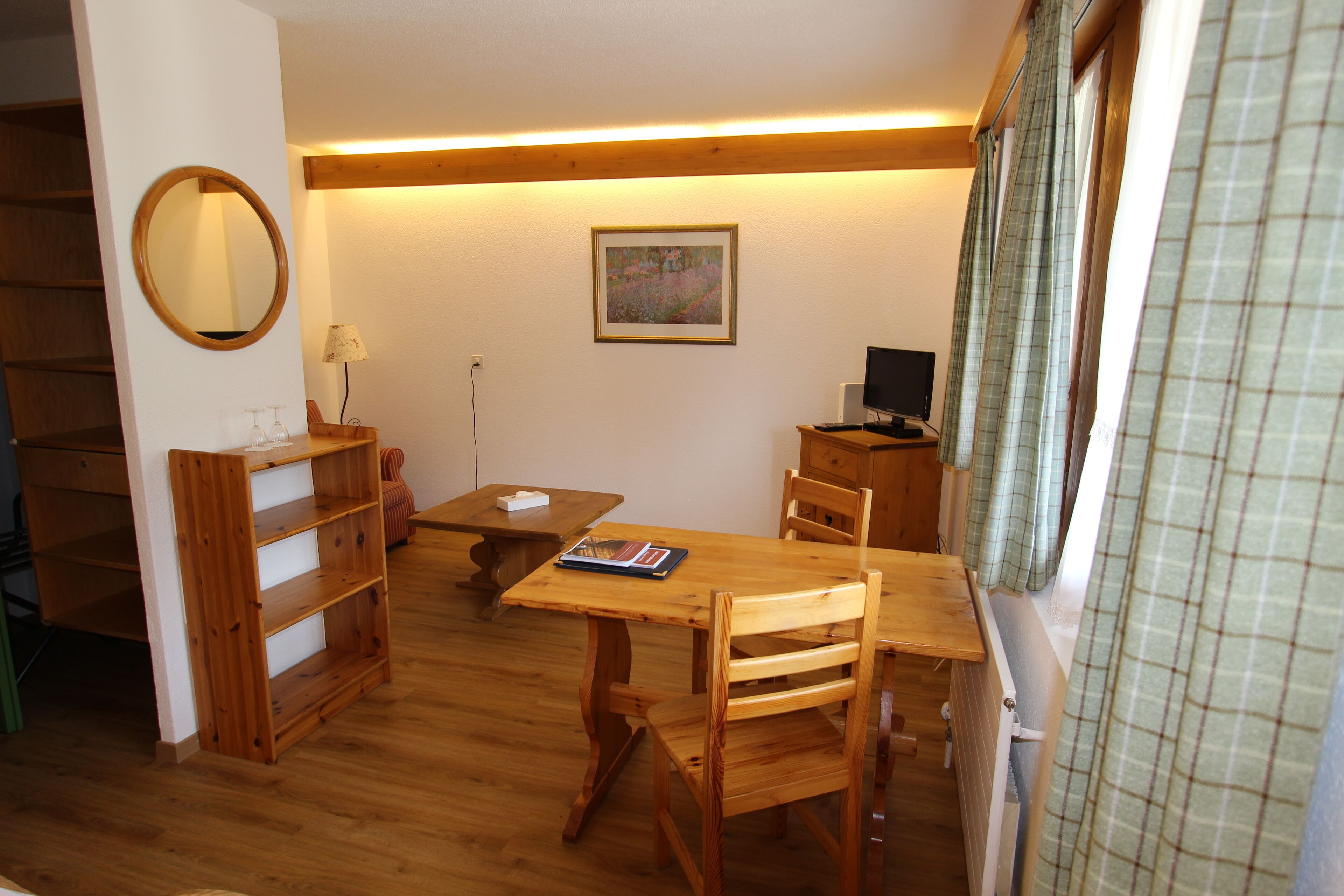 Una mesa de comedor de madera con sillas, una estantería de madera, un espejo, un cuadro en la pared y un televisor sobre un mueble de madera.