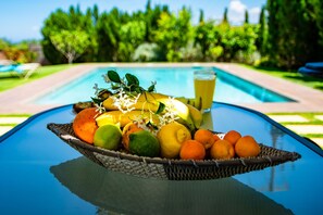 Un cistell de fruita sobre una taula amb una piscina i vegetació al fons.