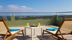 A balcony with two wooden lounge chairs, a small table with a plate of food and two glasses of orange juice, and a view of a coastal landscape. - Villa Dimokratia (Platanias)