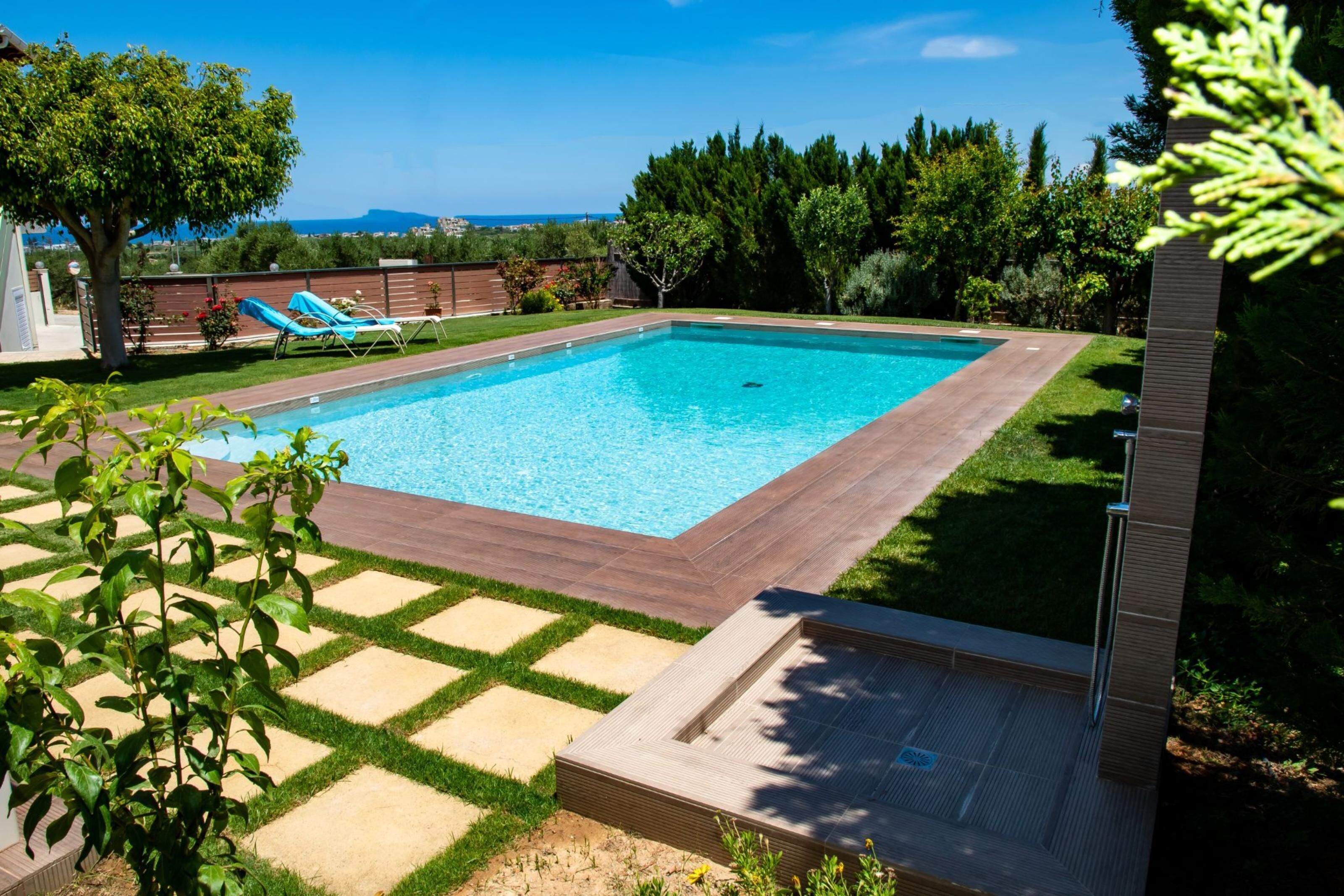 A rectangular swimming pool with a wooden deck and a tiled border, surrounded by a grassy lawn and trees.