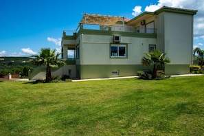 Une maison moderne à deux étages, dotée d’un balcon, entourée d’une pelouse bien entretenue et de palmiers.