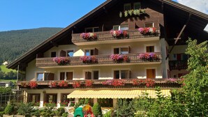 A multi-story building with balconies adorned with flowers, a covered outdoor seating area, and a mountainous backdrop. - Hotel Rodes (Castelrotto)