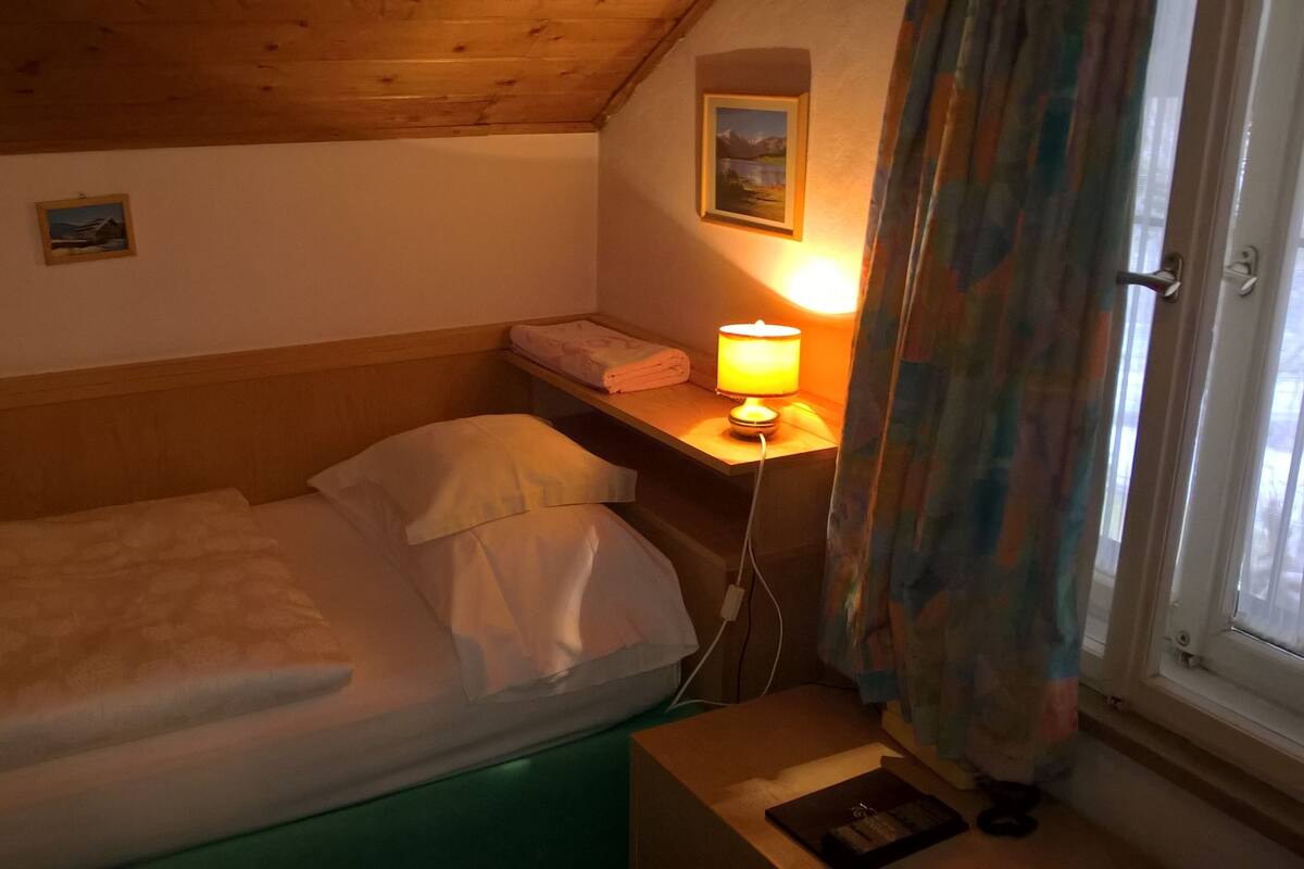 Una habitación pequeña y acogedora con una cama individual, una mesita de noche con lámpara y una ventana con cortinas.