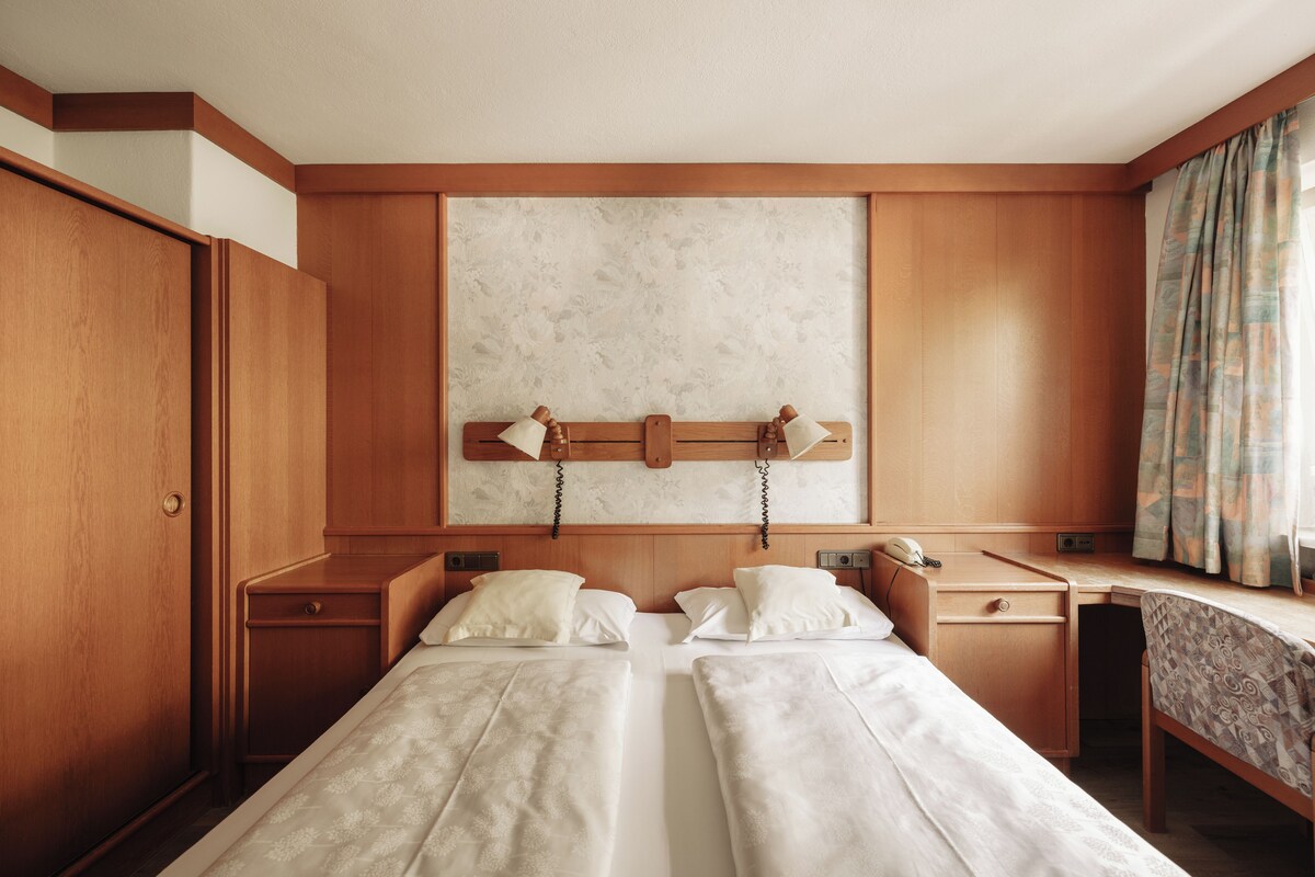 A hotel room with two single beds, a wooden desk, a telephone, and a window with curtains.