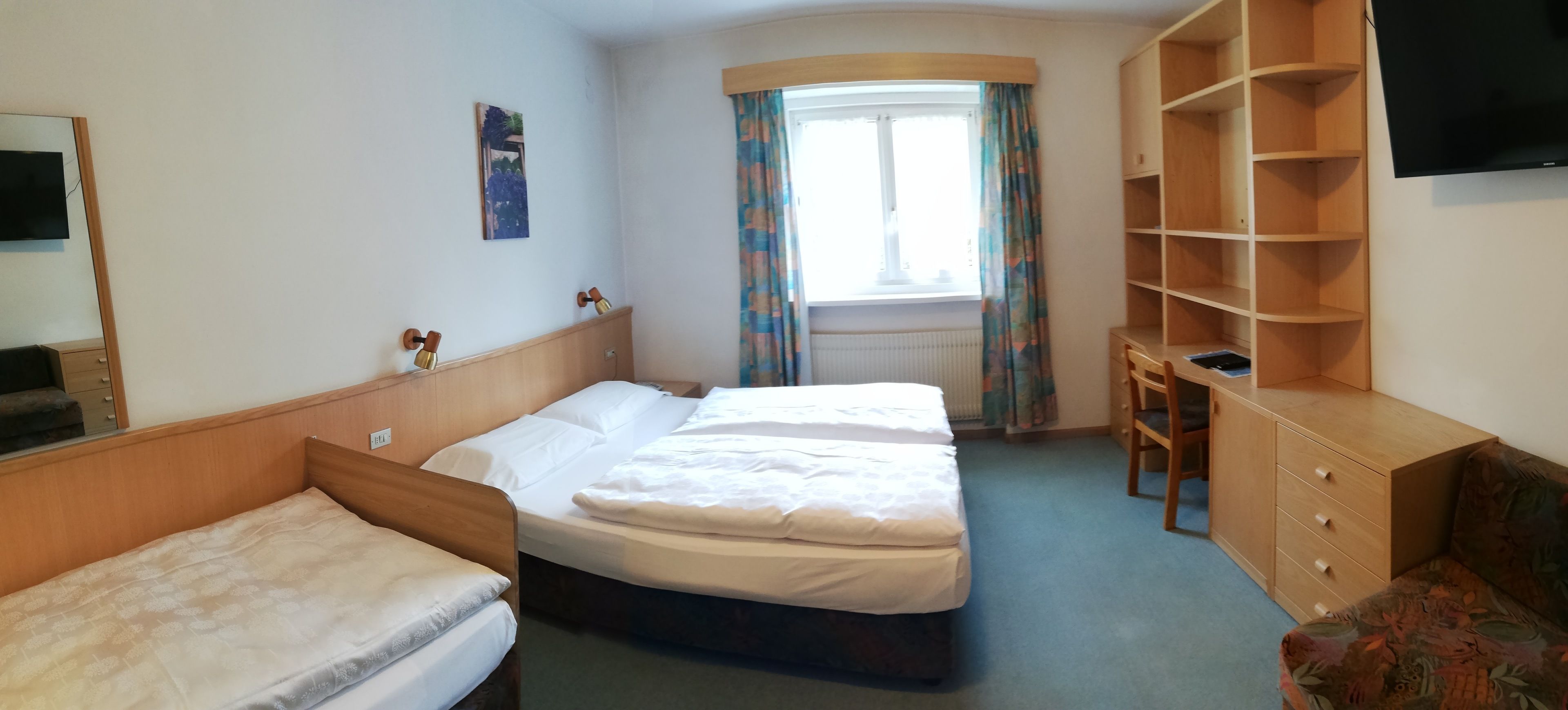 Ein Hotelzimmer mit einem Doppelbett, einem Einzelbett, einem Schreibtisch, einem Stuhl, einem Fernseher und einem Fenster mit Vorhängen.