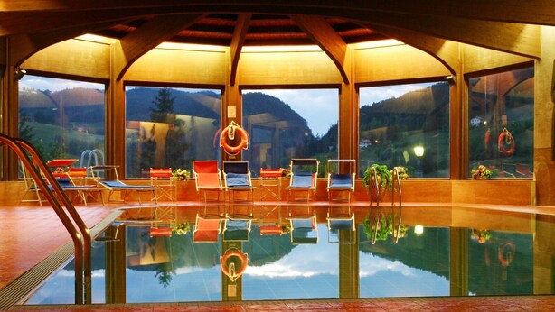 An indoor swimming pool with clear glass walls, multiple lounge chairs, and a view of a mountainous landscape. - Hotel Rodes (Castelrotto)