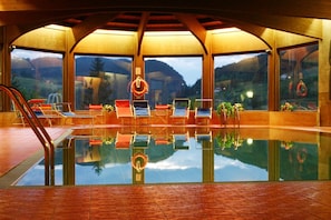 An indoor swimming pool with clear glass walls, multiple lounge chairs, and a view of a mountainous landscape.