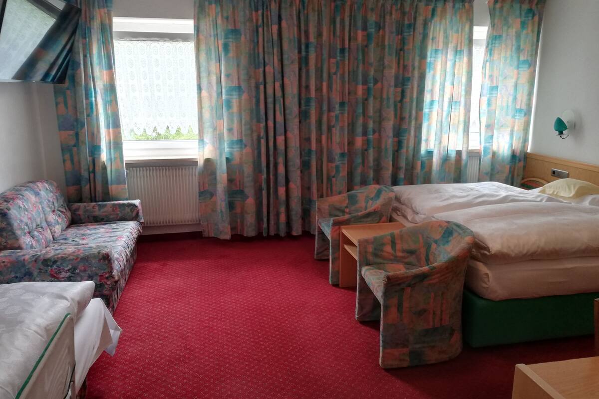Una habitación de hotel con alfombra roja, un sofá, una cama y una ventana con cortinas estampadas.