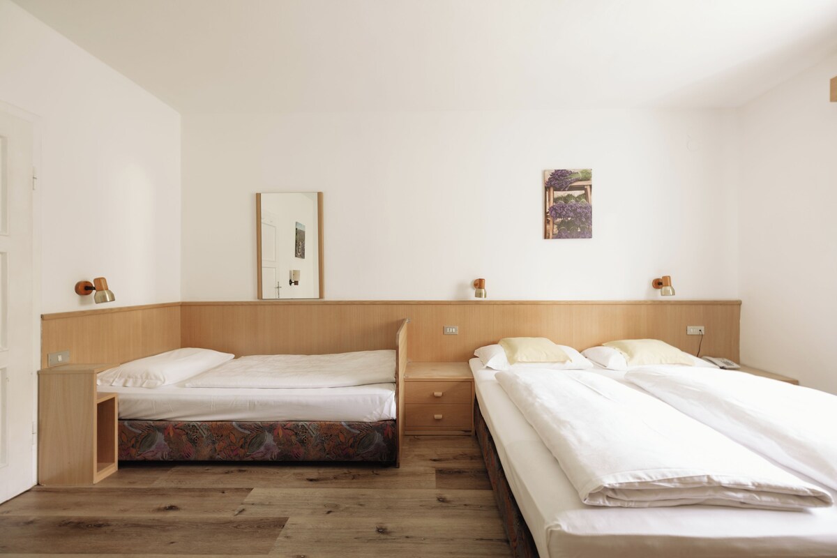 A hotel room with two beds, a wooden headboard, a small bedside table, a nightstand, a wall-mounted lamp, and a framed picture.