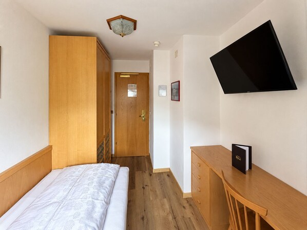 A hotel room with a bed, a desk, a television mounted on the wall, and a wooden wardrobe.