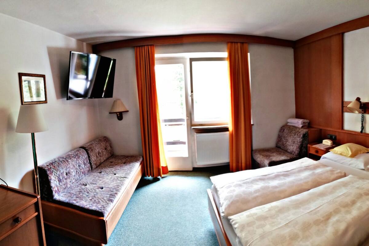 Habitación de hotel con sofá, dos camas individuales, televisor, lámpara y ventana con cortinas.