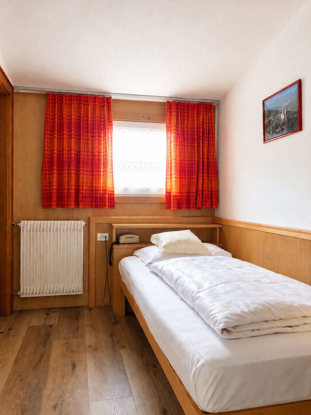 Habitación individual con una cama y cabecera de madera, una ventana con cortinas rojas, un radiador, un teléfono y un cuadro en la pared.