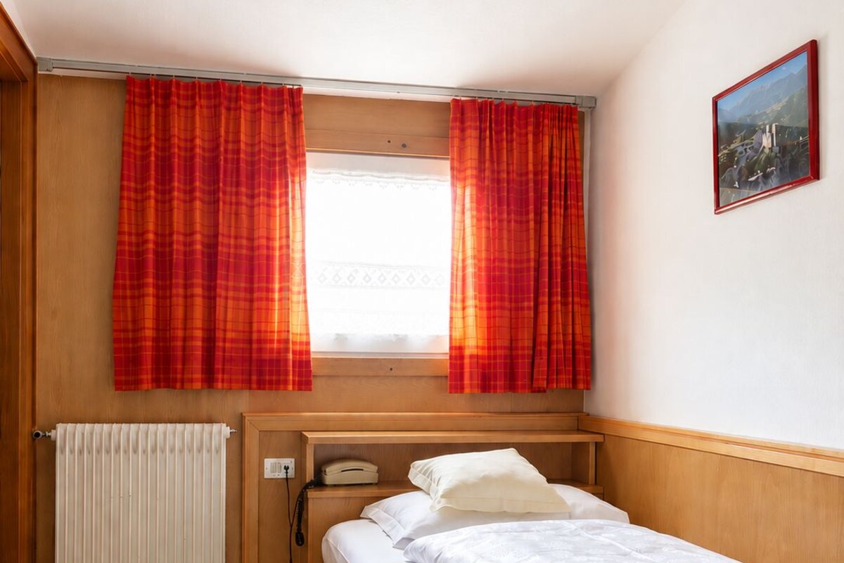 A single-bed room with a wooden headboard, a window with red curtains, a radiator, a telephone, and a framed picture on the wall.