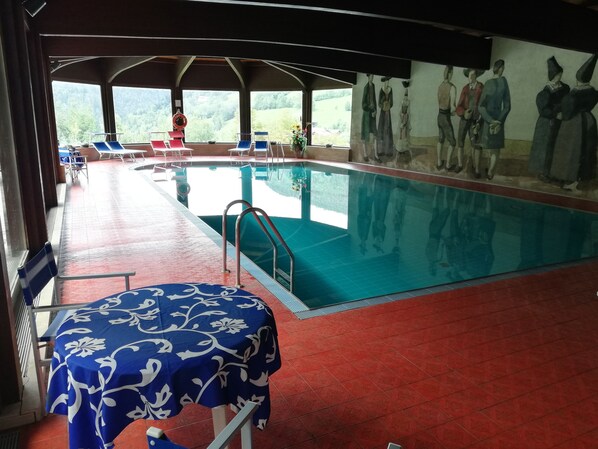 An indoor swimming pool with a mural, lounge chairs, and a table with a blue floral tablecloth.