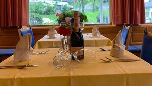 A dining table set with a bottle of Moët & Chandon champagne, a vase with flowers, and empty wine glasses. The table is covered with a yellow tablecloth, and there are blue cushioned seats. The room has large windows with red curtains, offering a view of a house and greenery outside. - Hotel Rodes (Castelrotto)