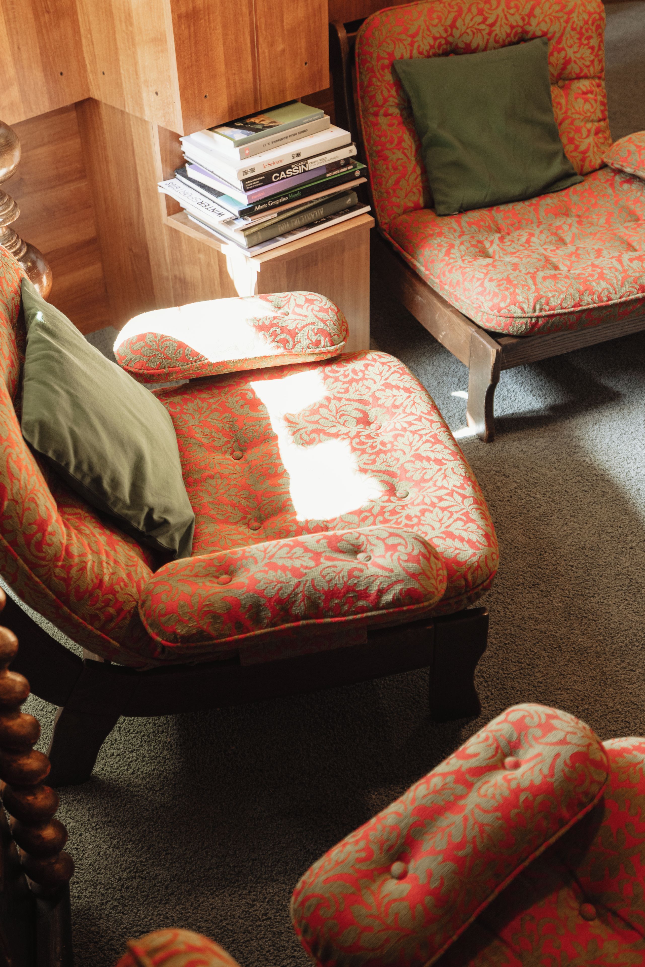 Una habitación con un sillón estampado, un otoman a juego y una pila de libros sobre una mesita de madera.