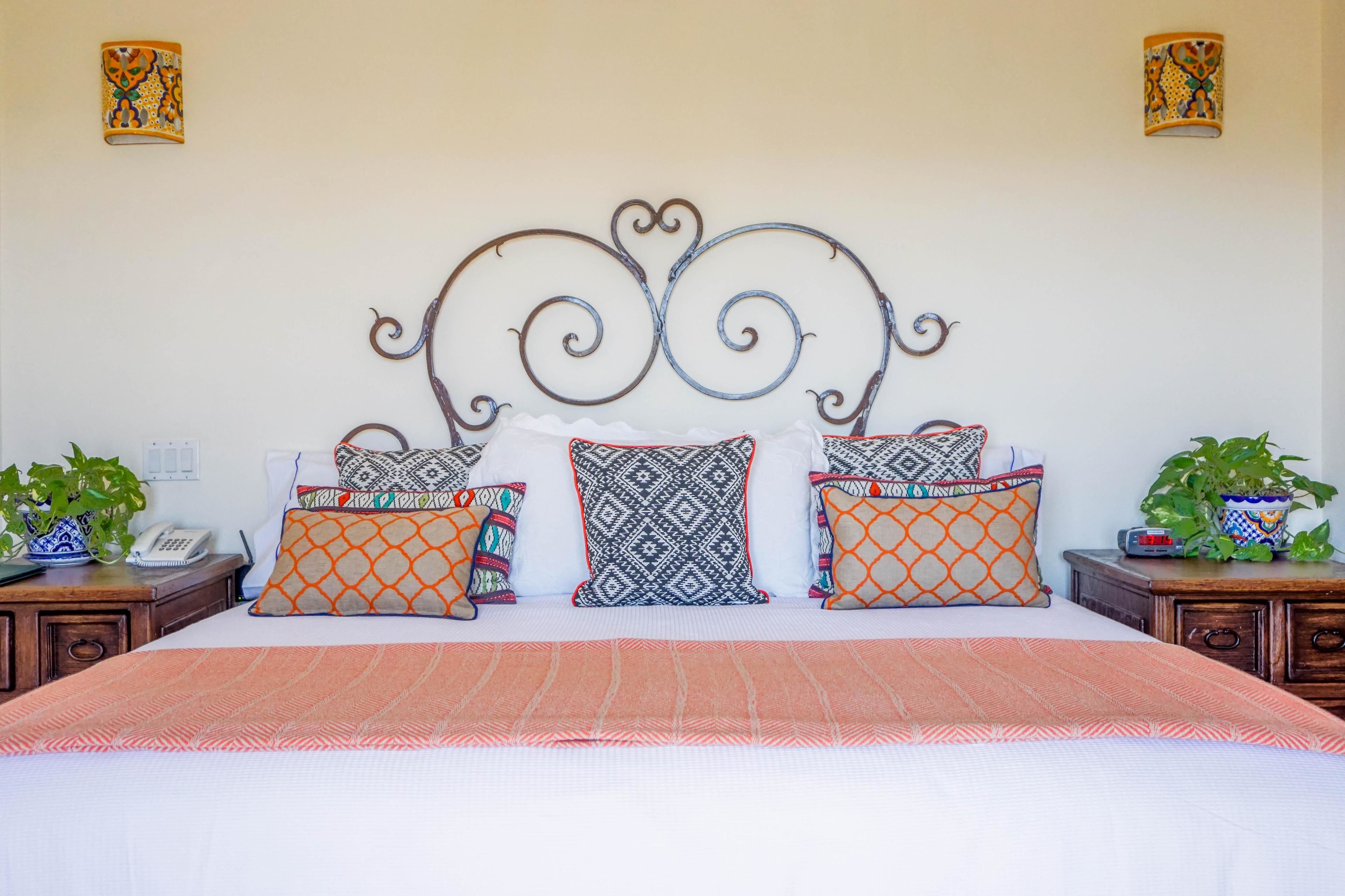 A neatly made bed with patterned pillows, a decorative metal headboard, and bedside tables with plants and a phone.