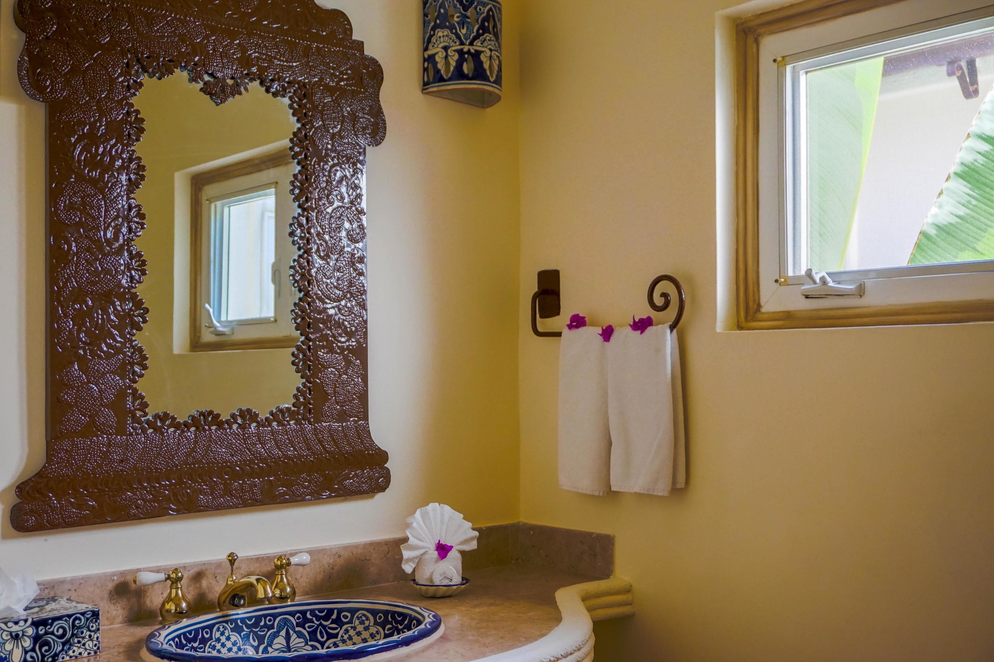 A bathroom with a large ornate mirror, a sink with decorative faucets, a towel rack with towels, and a window with a view of greenery.