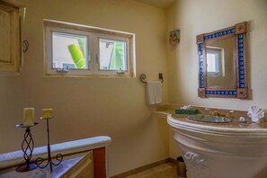 Ein Badezimmer mit Fenster, einem Waschbecken mit dekorativem Spiegel und zwei Kerzen auf einem Sockel.