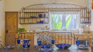 A kitchen with a wooden door, a window with a view of green foliage, and a metal rack with blue dishes.
