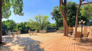 An outdoor wooden deck with tables and chairs, surrounded by trees and a stone wall. - Riverfront Bliss (New Braunfels)