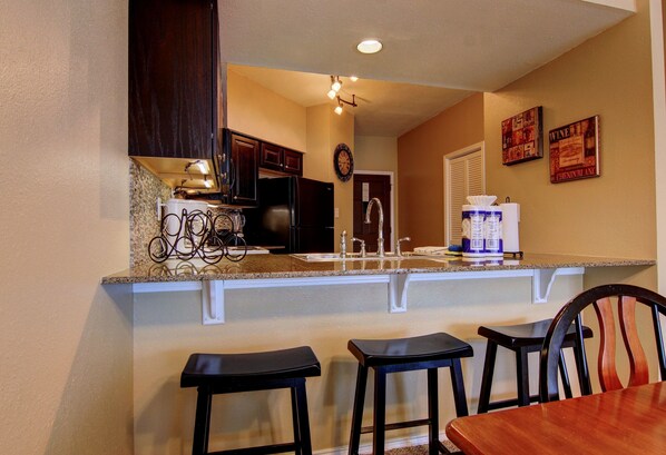 A kitchen with dark wooden cabinets, a granite countertop, a refrigerator, a sink, and a clock on the wall. There are two bar stools and a wooden table. - Riverfront Bliss (New Braunfels)