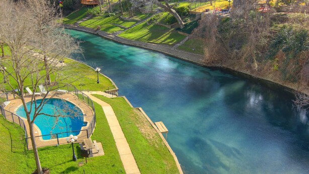 A swimming pool with a surrounding deck, a grassy area, and a river. - Riverfront Bliss (New Braunfels)