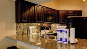 A kitchen with dark wooden cabinets, a granite countertop, a stainless steel sink, a black microwave, a black refrigerator, and a coffee maker. - Riverfront Bliss (New Braunfels)