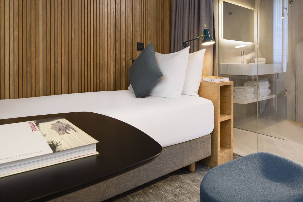 A modern hotel room with a neatly made bed, a bedside table with a lamp, a glass shower enclosure, and a wooden wall paneling.