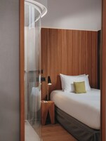 A modern hotel room with a wooden panel wall, a bed with white linens, a green pillow, a bedside table with a lamp, and a glass shower enclosure.