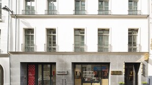 Un bâtiment à la façade moderne, avec un magasin au rez-de-chaussée doté de grandes vitrines et d’une enseigne indiquant « Atelier de dessin ».