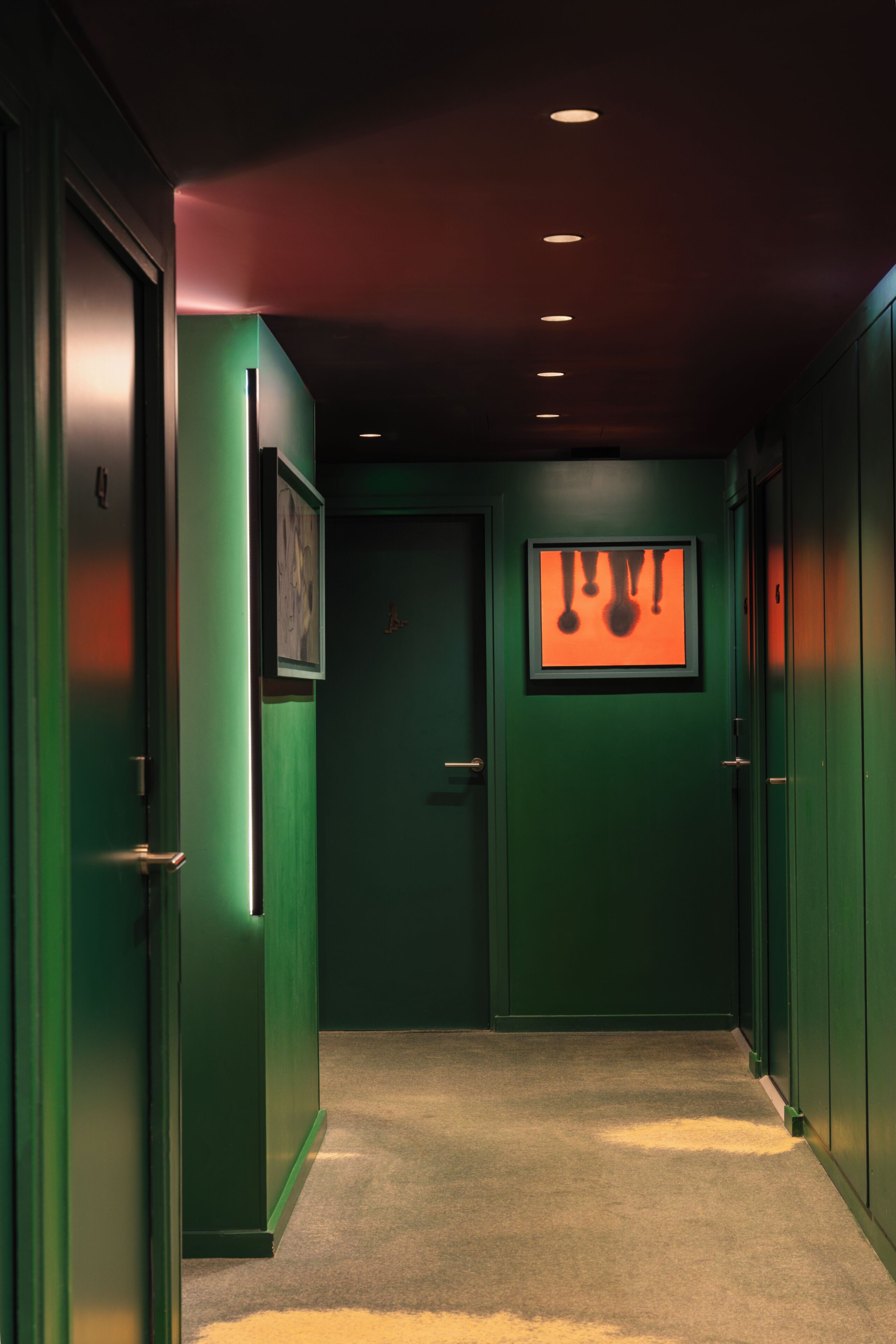 A hallway with green walls, a framed picture on the left, and a door with a red light.
