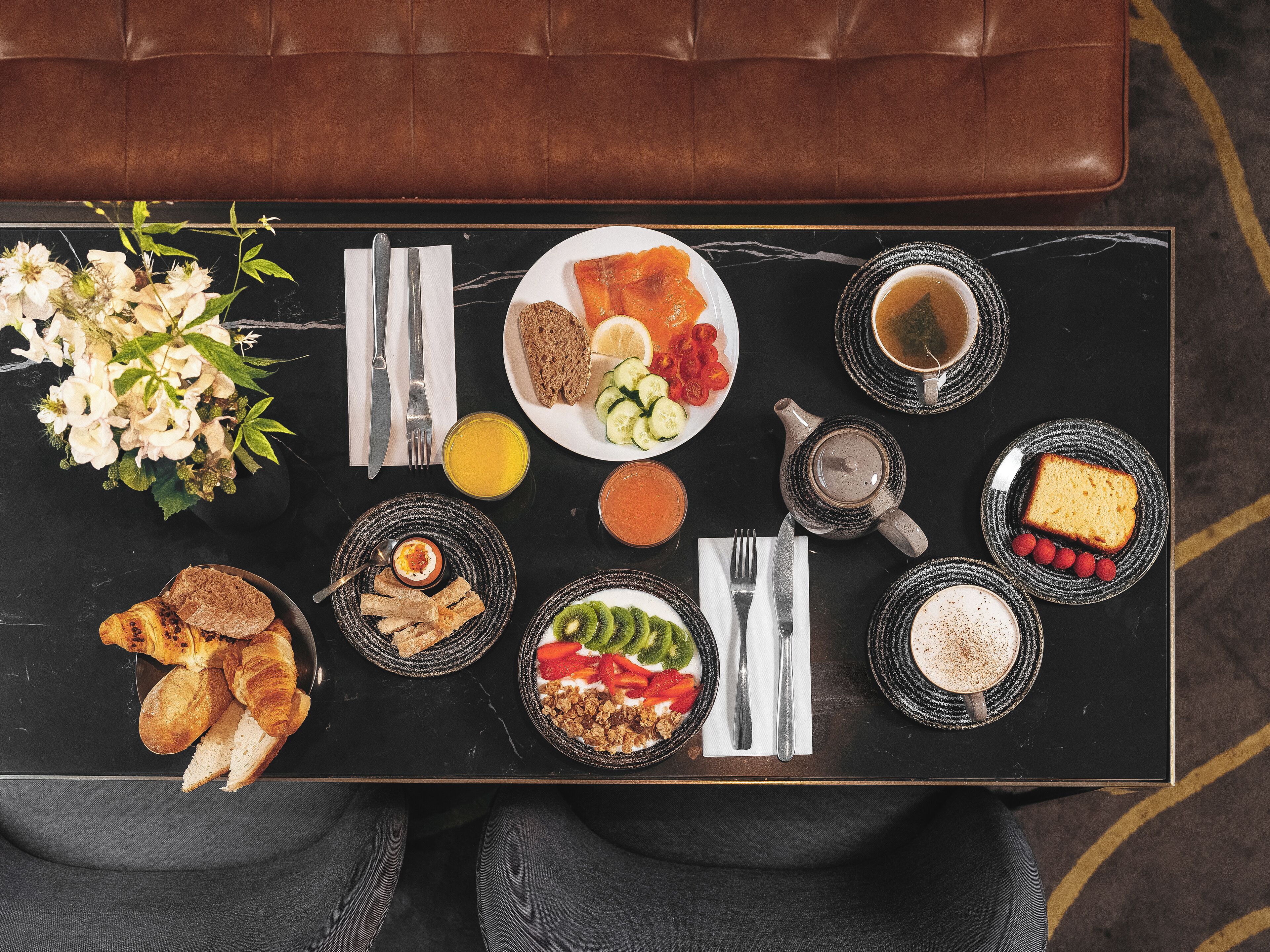 Een ontbijtplateau met croissants, fruit, yoghurt en koffie op een zwarte tafel.