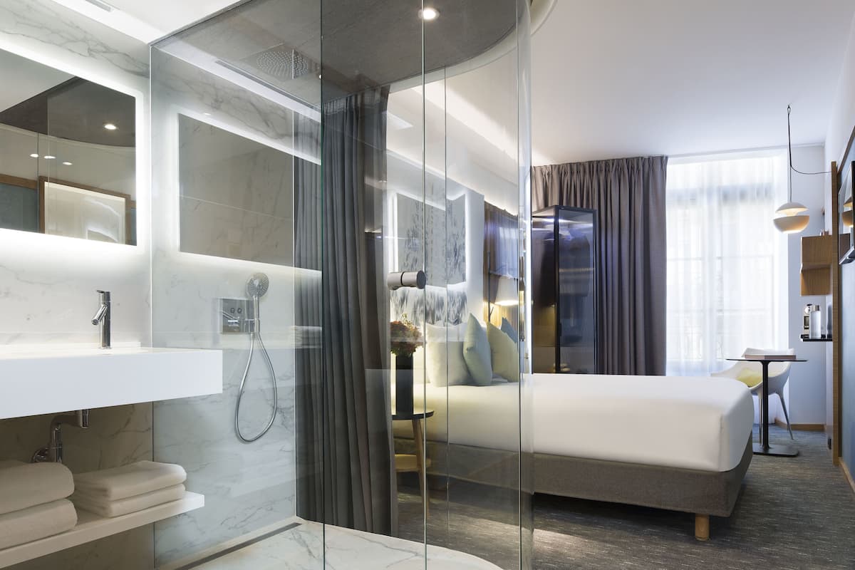 A modern hotel room with a glass-enclosed shower, a white bed, a small table, and a sofa.