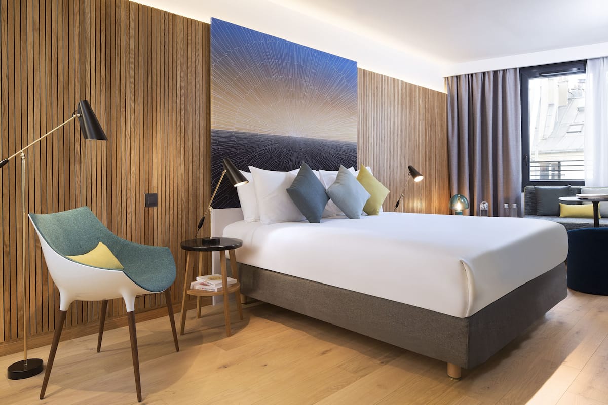 A modern hotel room with a large bed, a chair, a small table, and a large window.