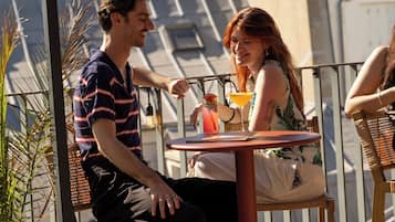 Un hombre y una mujer sentados en una mesa al aire libre, con bebidas, contemplando un paisaje urbano con edificios y cielo despejado.