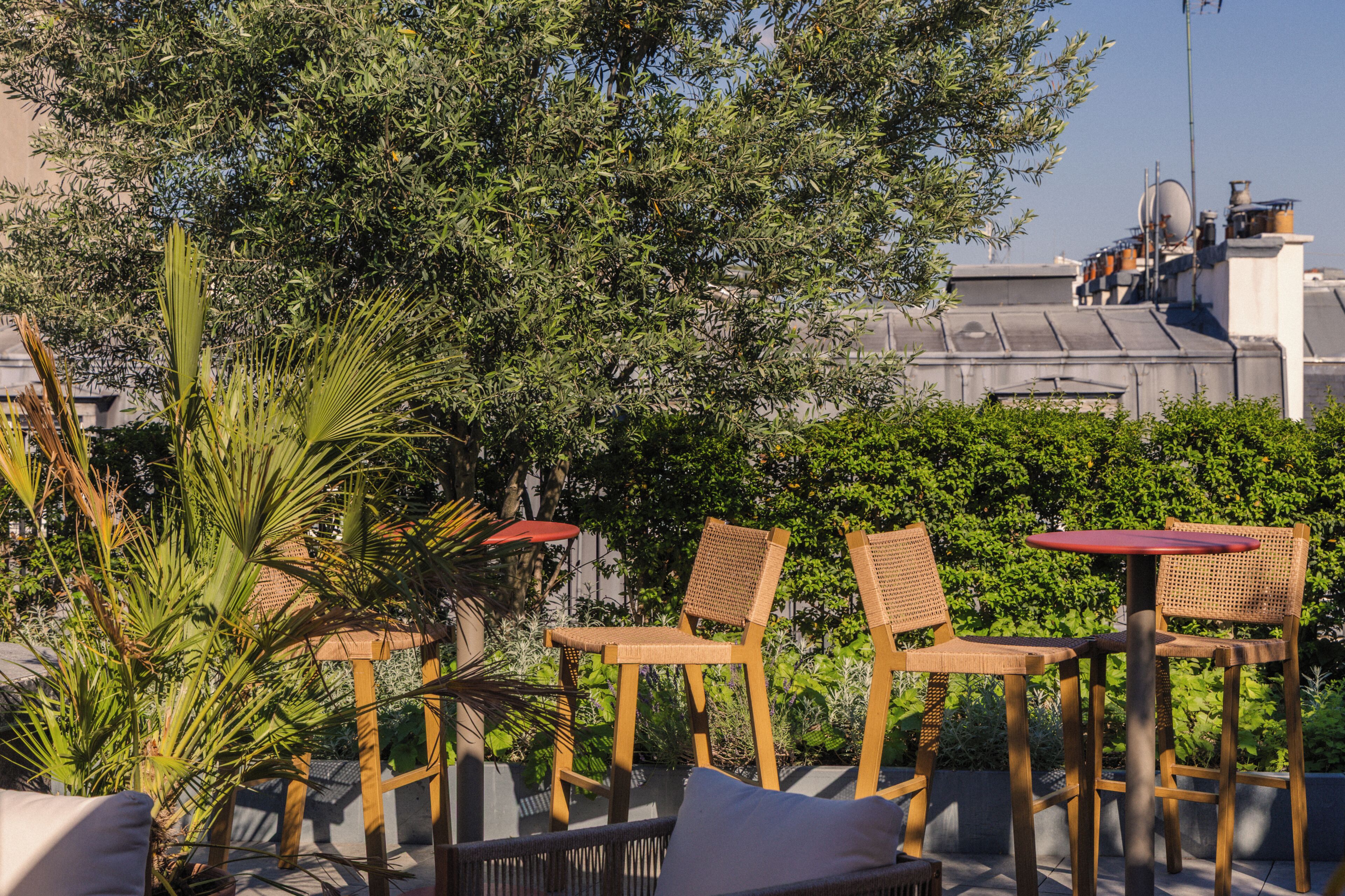 Outdoor seating with wicker chairs and a red table, surrounded by greenery and buildings in the background.