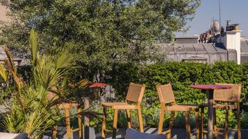 Outdoor seating with wicker chairs and a red table, surrounded by greenery and buildings in the background.