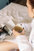 Uma pessoa sentada na cama com um livro e uma xícara.