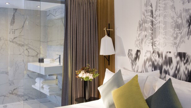 Une chambre d’hôtel moderne dotée d’une douche à parois de verre, d’une table de chevet blanche avec des articles de toilette, d’une lampe fixée au mur et d’un lit agrémenté de plusieurs oreillers.